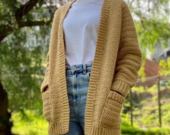 May include: A tan crocheted cardigan with pockets and a long sleeve. The cardigan is open in the front and is being worn over a white t-shirt and blue jeans.