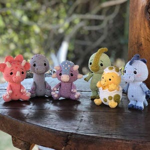 May include: A collection of six crocheted dinosaur toys in various colors. The dinosaurs include a pink triceratops, a gray dinosaur, a purple dinosaur, a green dinosaur, a yellow dinosaur in an egg, and a blue dinosaur.