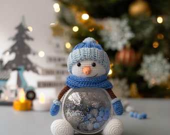 May include: A handmade snowman ornament with a blue and white knit hat, scarf, and mittens. The snowman has a clear glass body filled with silver glitter and blue beads. The snowman is sitting on a gray surface with a Christmas tree in the background.