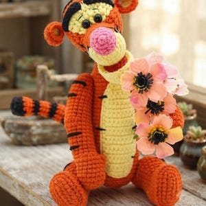 May include: A crocheted tiger figurine featuring orange and yellow yarn with black stripes, a pink nose, and black plastic eyes. The tiger is holding a small bouquet of three artificial light pink flowers with dark centers in its right paw.