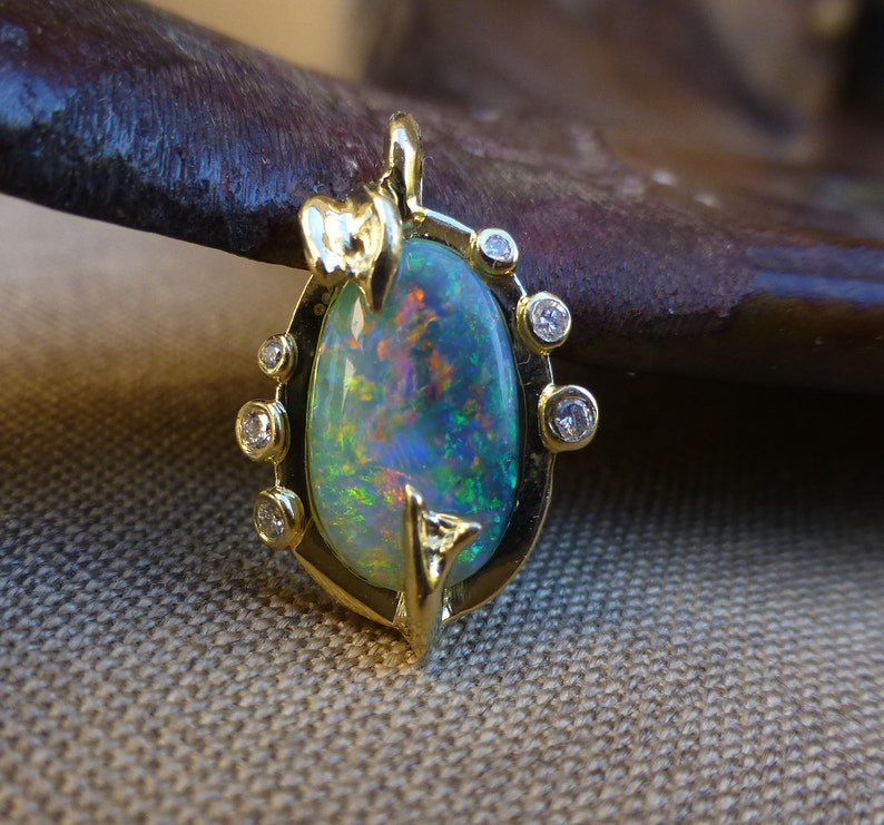 May include: A gold pendant featuring a large oval opal with a green, blue, and red iridescent sheen. The opal is surrounded by a gold setting with small diamonds.