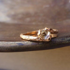 May include: A gold ring with a textured band and a clear gemstone. The ring is set on a brown surface.
