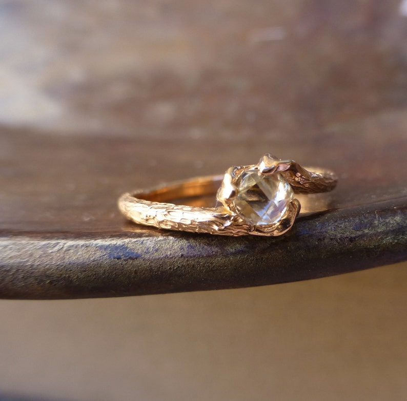 May include: A gold ring with a textured band and a clear gemstone. The ring is set on a brown surface.