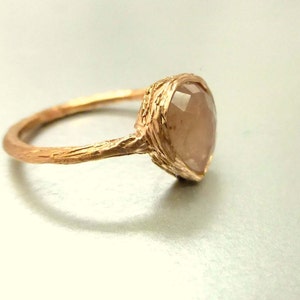 May include: A rose gold ring with a textured band and a large, faceted, light pink gemstone.