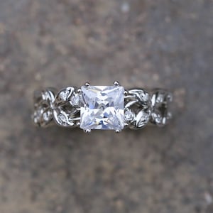 May include: A silver ring with a large square-shaped clear gemstone surrounded by smaller clear gemstones and a leaf design.
