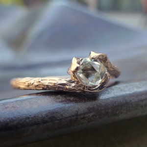 May include: A gold ring with a textured band and a clear, faceted gemstone. The ring is designed to look like a branch with a gemstone set in the center.