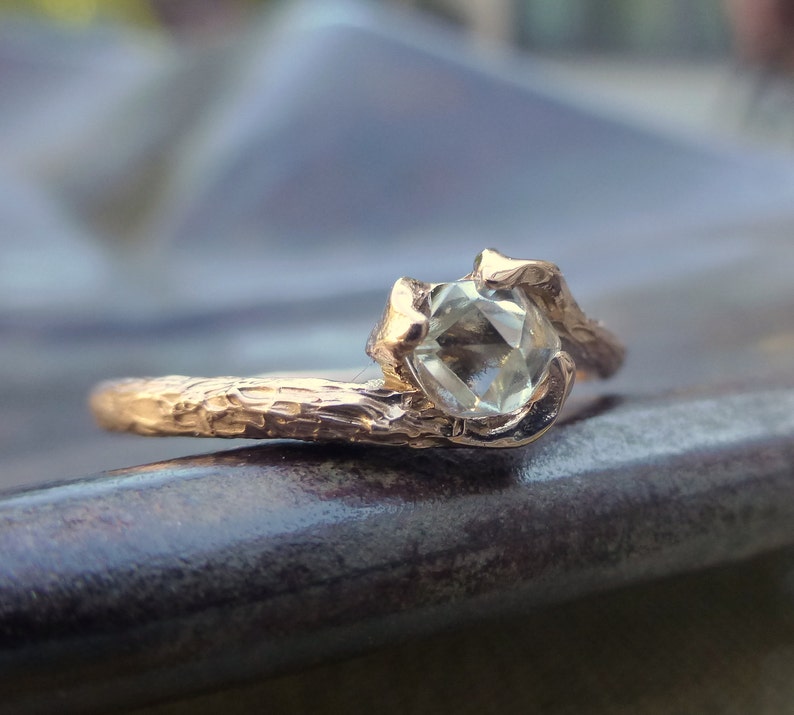 May include: A gold ring with a textured band and a clear, faceted gemstone. The ring is designed to look like a branch with a gemstone set in the center.