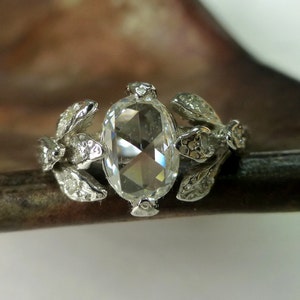 May include: A silver ring with a large, clear, oval-shaped gemstone surrounded by smaller gemstones. The ring band is decorated with delicate, leaf-like designs.