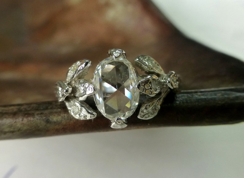 May include: A silver ring with a large, clear, oval-shaped gemstone surrounded by smaller gemstones. The ring band is decorated with delicate, leaf-like designs.