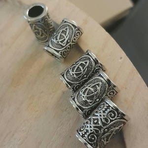May include: A set of five cylindrical metal beads featuring an antique silver finish and intricate embossed triquetra knotwork patterns. Each bead has a hollow center and detailed scrollwork surrounding the central triangular knot design.
