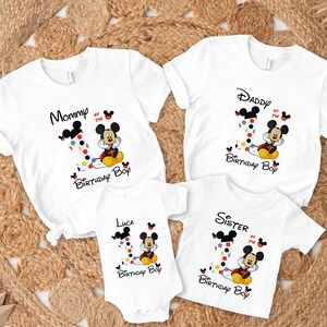 以下が含まれることがあります： ミッキーマウスのグラフィックが描かれた白いTシャツとボディスーツ。Tシャツには「Mommy」、「Daddy」、「Sister」と下に「Birthday Boy」と書かれています。ボディスーツには「Luca」と書かれています。各デザインには、水玉模様の数字の1が含まれています。