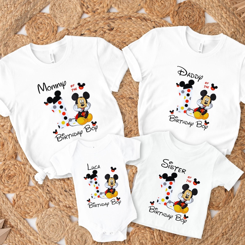以下が含まれることがあります： ミッキーマウスのグラフィックが描かれた白いTシャツとボディスーツ。Tシャツには「Mommy」、「Daddy」、「Sister」と下に「Birthday Boy」と書かれています。ボディスーツには「Luca」と書かれています。各デザインには、水玉模様の数字の1が含まれています。