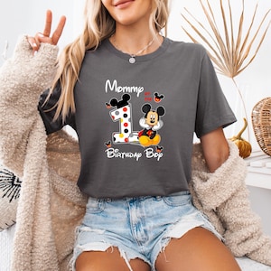 以下が含まれることがあります： 「Mommy of the Birthday Boy」の文字が入った濃いグレーのTシャツ。ミッキーマウスと数字の1のグラフィックが特徴で、水玉模様とミッキーマウスのシルエットで装飾されています。Tシャツはデニムショーツと組み合わせています。