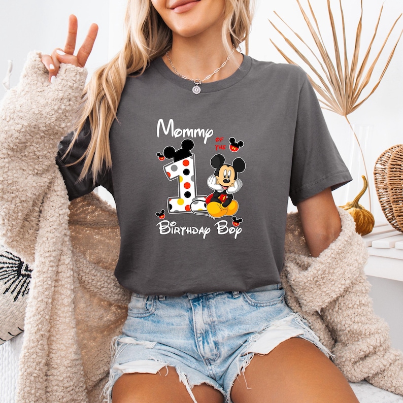 以下が含まれることがあります： 「Mommy of the Birthday Boy」の文字が入った濃いグレーのTシャツ。ミッキーマウスと数字の1のグラフィックが特徴で、水玉模様とミッキーマウスのシルエットで装飾されています。Tシャツはデニムショーツと組み合わせています。
