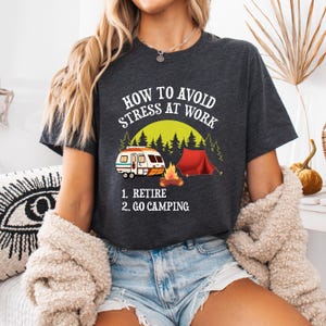 May include: Dark gray t-shirt with the text "HOW TO AVOID STRESS AT WORK." The graphic includes a camper, a red tent, a campfire, and trees. Below the graphic are the words "1. RETIRE 2. GO CAMPING."