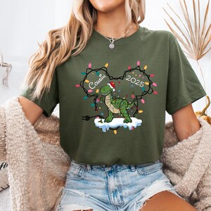 May include: Olive green t-shirt featuring a cartoon dinosaur wearing a Santa hat and wrapped in Christmas lights. The text "Cousin 2025" is above the dinosaur. The dinosaur is standing on a cloud of snow.