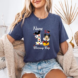 以下が含まれることがあります： ミッキーマウスと数字1のグラフィックが特徴のネイビーブルーのTシャツ。「Name」の文字が数字の上に、「Birthday Boy」が下にあります。デザインにはミッキーマウスの耳が含まれています。楽しくカラフルなデザインです。
