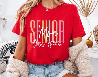 Op de afbeelding: Een rood t-shirt met de woorden "SENIOR Mom Class of 2026" in een kleurrijk ontwerp. Het shirt heeft korte mouwen en is gemaakt van een zacht materiaal. De tekst is in een mix van lettertypen en kleuren.