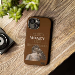 Könnte beinhalten: Braune Handyhülle mit dem Text "NEED MONEY NOT BOYFRIEND" in Weiß. Die Hülle hat ein Rasterdesign und ein Graustufenbild einer Skulptur. Die Hülle liegt auf einer Holzoberfläche.