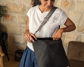 May include: A woman is holding a black canvas tote bag with a grey leather strap. The bag is worn over the shoulder and has a simple design.