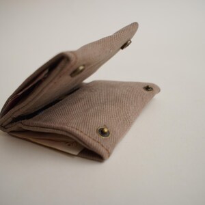 May include: A light brown canvas wallet, open to reveal the interior. The wallet has a rectangular shape with rounded corners and is secured with brass snap closures. The fabric has a textured appearance.