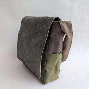 May include: A green, brown, and beige messenger bag with a flap closure. The bag has a textured, leather-like flap and a canvas body.