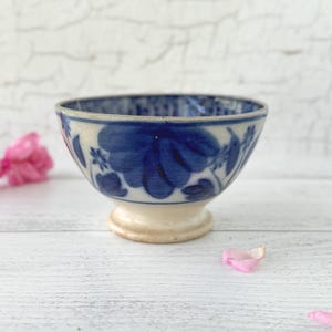 May include: A small, antique-style ceramic bowl with a white base and a blue floral pattern. The bowl has a slightly flared rim and is decorated with stylized blue flowers and leaves. The bowl is set against a white background with pink flower petals.