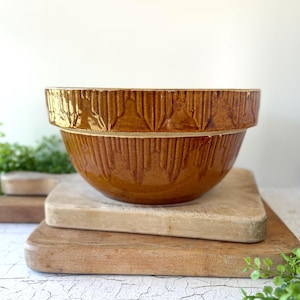 May include: A round, glazed ceramic mixing bowl in a deep amber-brown color. The exterior features a repeating vertical ribbed pattern around the upper rim and body, with a smooth, rounded base and a wide, open top opening.