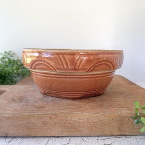 May include: A round, glazed ceramic bowl in a warm terracotta brown color. The exterior features a decorative embossed pattern consisting of repeating arched lines and vertical segments encircling the upper portion of the bowl's side.