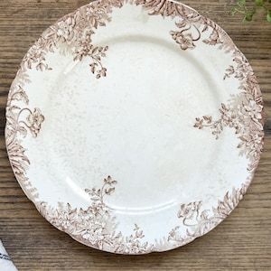 May include: A round ceramic dinner plate with a cream-colored base and a scalloped edge. The rim is decorated with a detailed brown floral pattern featuring leaves and blossoms that extend slightly toward the center of the plate.