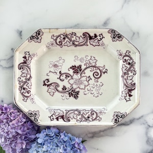 May include: An antique, octagonal serving platter with a cream-colored background and a floral design in burgundy. The edges of the platter are also burgundy. The floral design includes flowers and swirling leaves. The platter is likely made of ceramic.