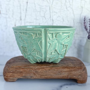 May include: A mint green ceramic bowl with a leaf design, resting on a small, rectangular wooden stand. The bowl's exterior features a repeating pattern of embossed leaves. The background is a white wall with a distressed, crackled paint effect.