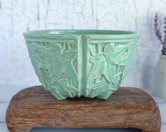 Pode incluir: Uma tigela de cerâmica verde menta com um desenho de folhas, repousando sobre um pequeno suporte de madeira retangular. O exterior da tigela apresenta um padrão repetido de folhas em relevo. O fundo é uma parede branca com efeito de pintura rachada.