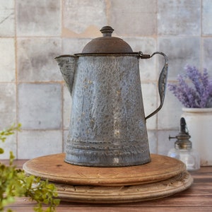 May include: Vintage gray enamelware coffee pot with a dark brown lid and handle. The pot has a speckled pattern and is sitting on a wooden cutting board. The background features a tiled wall and a vase of lavender.
