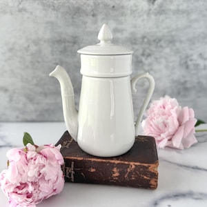 May include: A white ceramic coffee pot with a lid, spout, and handle. The pot is sitting on a dark brown book, with pink peonies in the foreground and background. The background is a light gray.