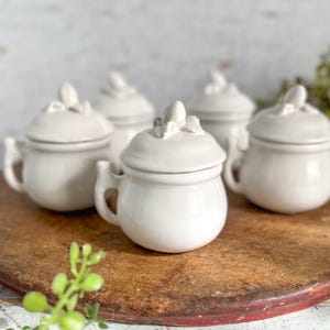 May include: A collection of five small, off-white ceramic containers with lids. Each container has a rounded body, a small handle, and a lid with a decorative floral accent. The containers are arranged on a rustic, wooden surface.