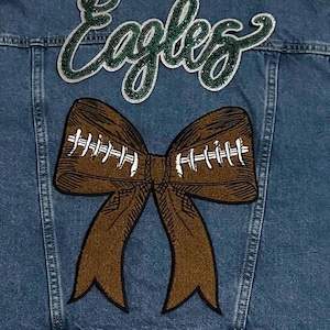 Op de afbeelding: Een spijkerjasje in een medium blauwe wassing met het woord "Eagles" in groene glitterletters op de rug. Daaronder een bruine, football-vormige strik met witte stiksels. Casual en sportief.
