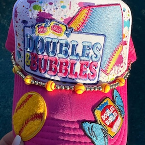 May include: Pink trucker hat with a white top featuring colorful paint splatters. The hat has the text "DOUBLES & BUBBLES" and a softball patch. A beaded necklace with yellow baseballs is attached to the brim.