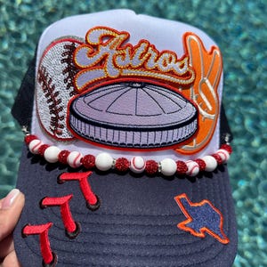 May include: A baseball cap with a white front panel and a navy blue brim. The cap features the word "Astros" in orange and white, a baseball, and a peace sign. The brim has red "T" patches and a Texas outline.