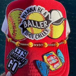 May include: Red trucker hat with white mesh backing, adorned with softball patches and a beaded chain. Patches include a softball with the text "Wanna Be a Baller Shot Caller", and "Let's Freaking Go".