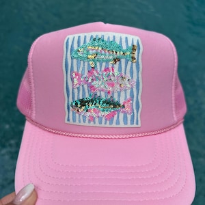 May include: A light pink trucker hat featuring a rectangular patch on the front panel. The patch displays three stacked fish designs embellished with colorful sequins in shades of teal, pink, and white, set against a background of vertical blue stripes.