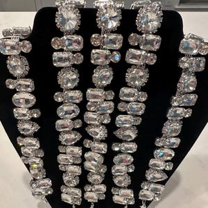 May include: A display of several silver-toned necklaces with large, clear, faceted crystal stones. The necklaces are arranged on a black velvet display stand, showcasing the varying shapes and sizes of the crystals.