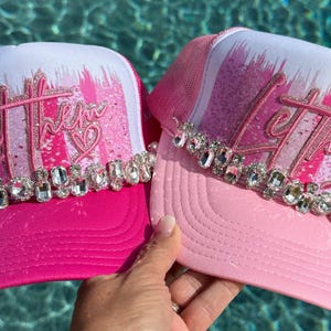 Op de afbeelding: Twee truckerpetten in roze en witte tinten, voorzien van een glinsterende roze cursieve tekst Let them met een klein hartje. Elke pet is versierd met een horizontale rij van heldere, gefacetteerde strass-steentjes op het voorpaneel.