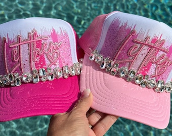 May include: Two trucker hats in shades of pink and white, featuring a glittery pink script text that reads Let them with a small heart icon. Each hat is adorned with a horizontal row of clear, faceted rhinestones across the front panel.