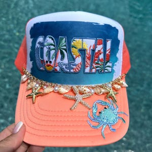 Op de afbeelding: Een perzikkleurige truckerpet met een wit voorpaneel waarop het woord COASTAL in het blauw is gedrukt, versierd met een goudkleurige ketting, kleine witte parels en twee zeesterbedels. Een lichtblauwe geborduurde krab zit op de klep.