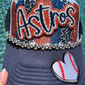 May include: A navy blue trucker hat with a white mesh back. The front features the word "Astros" in orange glitter, a rhinestone band, and a baseball heart patch. The hat has a patterned design in the background.