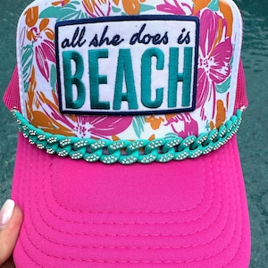May include: A bright pink trucker hat featuring a white front panel with a colorful floral print in shades of orange and pink. A rectangular patch displays the text all she does is BEACH in teal. A teal chain with rhinestones accents the brim.