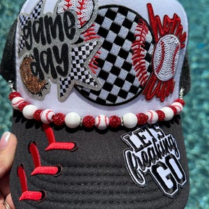 May include: Baseball-themed trucker hat with a white front panel and black mesh back. Embellished with baseball graphics, the words "Game Day" and "Home Run", and the phrase "Let's Freaking Go". A beaded baseball trim adds detail.