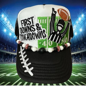 May include: Black and white trucker hat with a football theme. The hat features the text "FIRST DOWNS & TOUCHDOWNS" and an image of a referee signaling a touchdown. A beaded necklace with football and white beads adorns the hat.