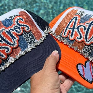 May include: Two trucker hats featuring the word Astros in cursive script. One hat is navy blue with a white front panel, and the other is orange with a white front panel. Both feature glittery leopard print accents, rhinestone trim, and a baseball heart patch.